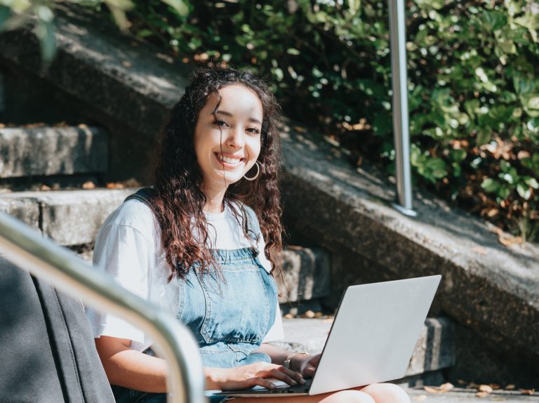 Side-shot enthusiastic happy carefree female student prepare project working laptop sitting outside the university wear modern outfit enjoy climate smiling to camera and looking the screen. Copy space