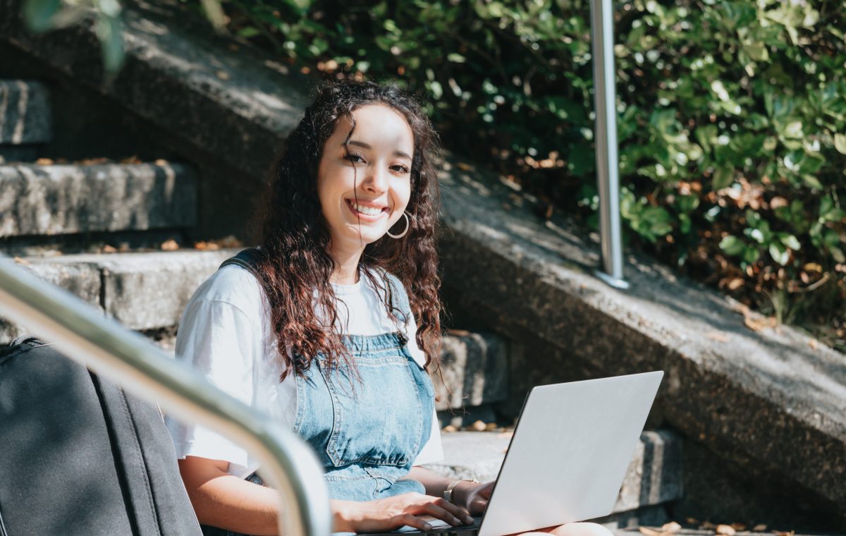 Side-shot enthusiastic happy carefree female student prepare project working laptop sitting outside the university wear modern outfit enjoy climate smiling to camera and looking the screen. Copy space